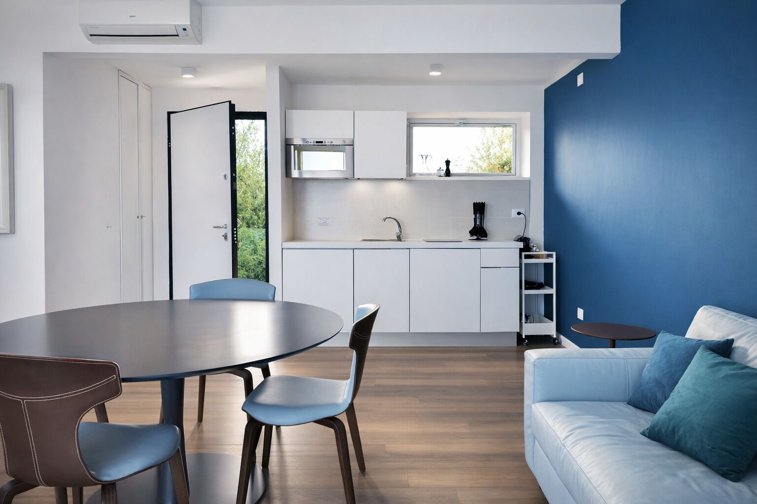 Una cocina moderna con armarios blancos, un microondas y un ventanal. Hay una zona de comedor con una mesa y sillas, además de un sofá azul con cojines.