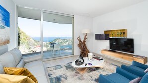 A modern living room with a sofa, a blue armchair, a coffee table, a TV, and a view of the sea through large glass doors. - Isola Bella Infinity Suites (Taormina)