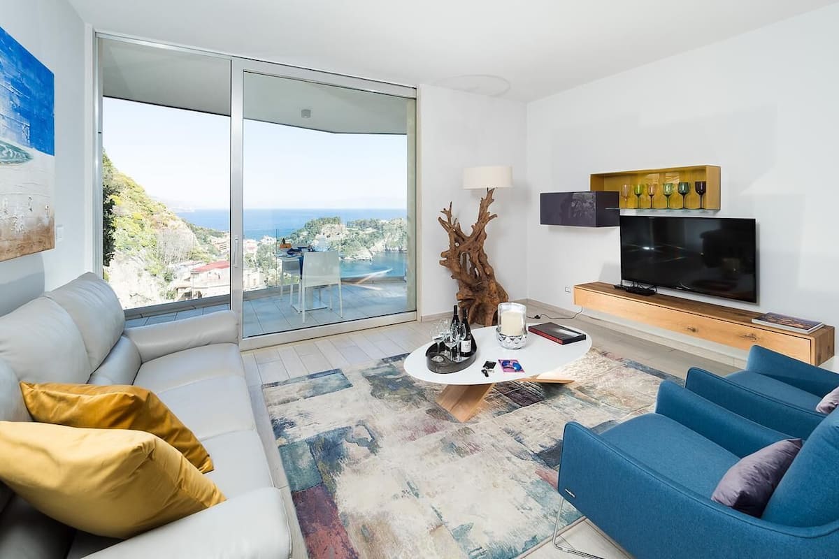 Una sala de estar moderna con un sofá, un sillón azul, una mesa de centro, un televisor y vistas al mar a través de puertas de cristal amplias.