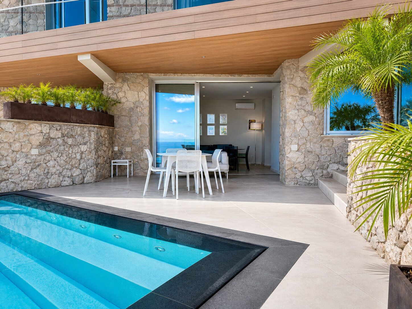 A modern outdoor pool area with a stone wall, a dining set, and a view of the ocean.