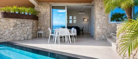 A modern outdoor pool area with a stone wall, a dining set, and a view of the ocean.