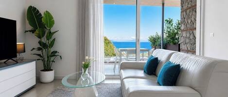 Ett modernt vardagsrum med en soffa, ett soffbord i glas, en tv och utsikt över havet.