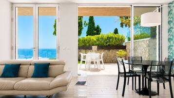 A modern living room with a sofa, a dining table with chairs, and a view of the sea through large sliding doors.