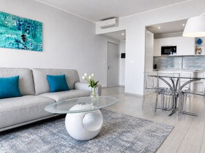 A modern living room with a glass coffee table, a sofa with blue cushions, a vase with flowers, and a kitchen area with a microwave and a dining set. - Isola Bella Infinity Suites (Taormina)