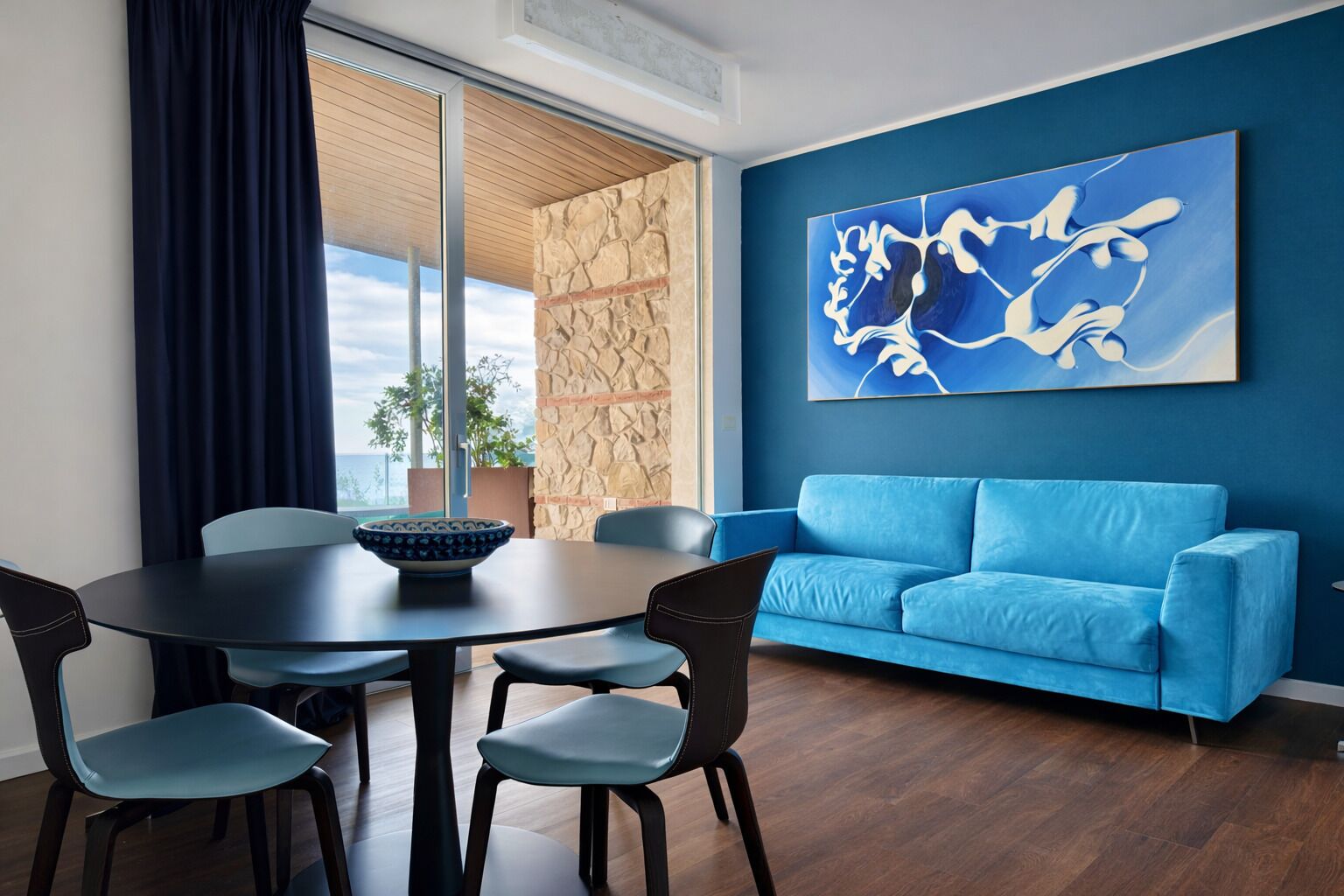 A modern living room with a blue sofa, a dining table with chairs, and a large abstract painting on the wall.