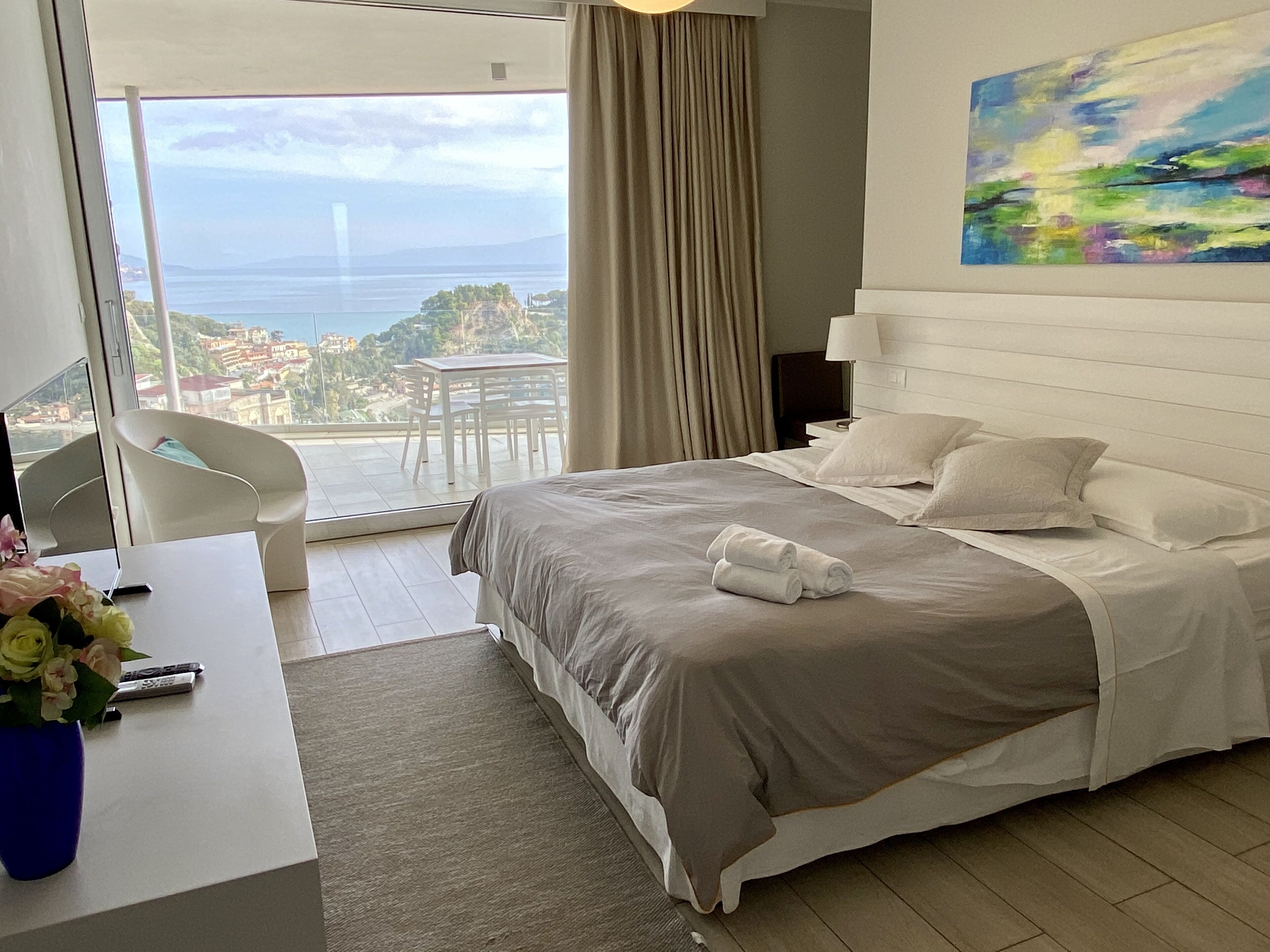 Une chambre d’hôtel avec un lit, un bureau agrémenté d’un vase de fleurs, une chaise, un tableau au mur et une vue sur la mer et les bâtiments.