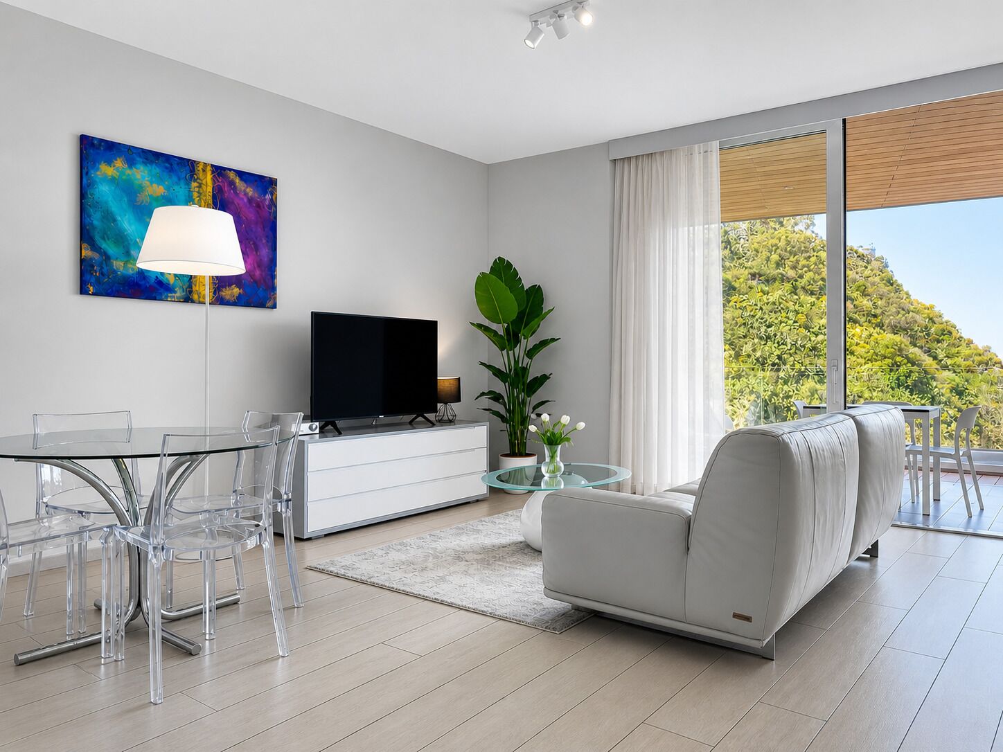 A modern living room with a flat-screen TV, a sofa, a dining table with chairs, and a large window with a view of greenery.