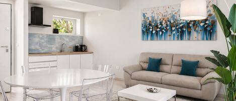 A modern living room with a white dining table, clear chairs, a beige sofa with teal cushions, a coffee table, and a large abstract painting on the wall.
