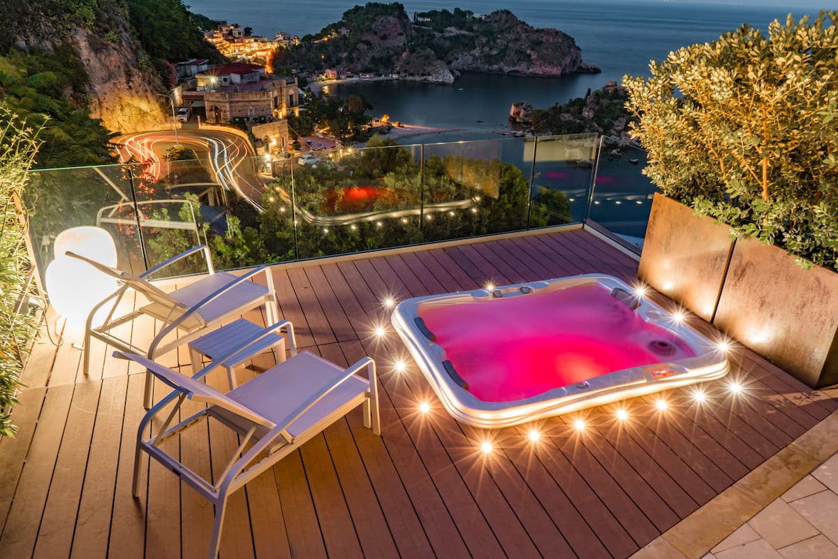 Terraza de madera con jacuzzi, sillas de descanso y vistas a un pueblo costero al atardecer.