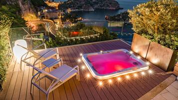 Terraza de madera con jacuzzi, sillas de descanso y vistas a un pueblo costero al atardecer.