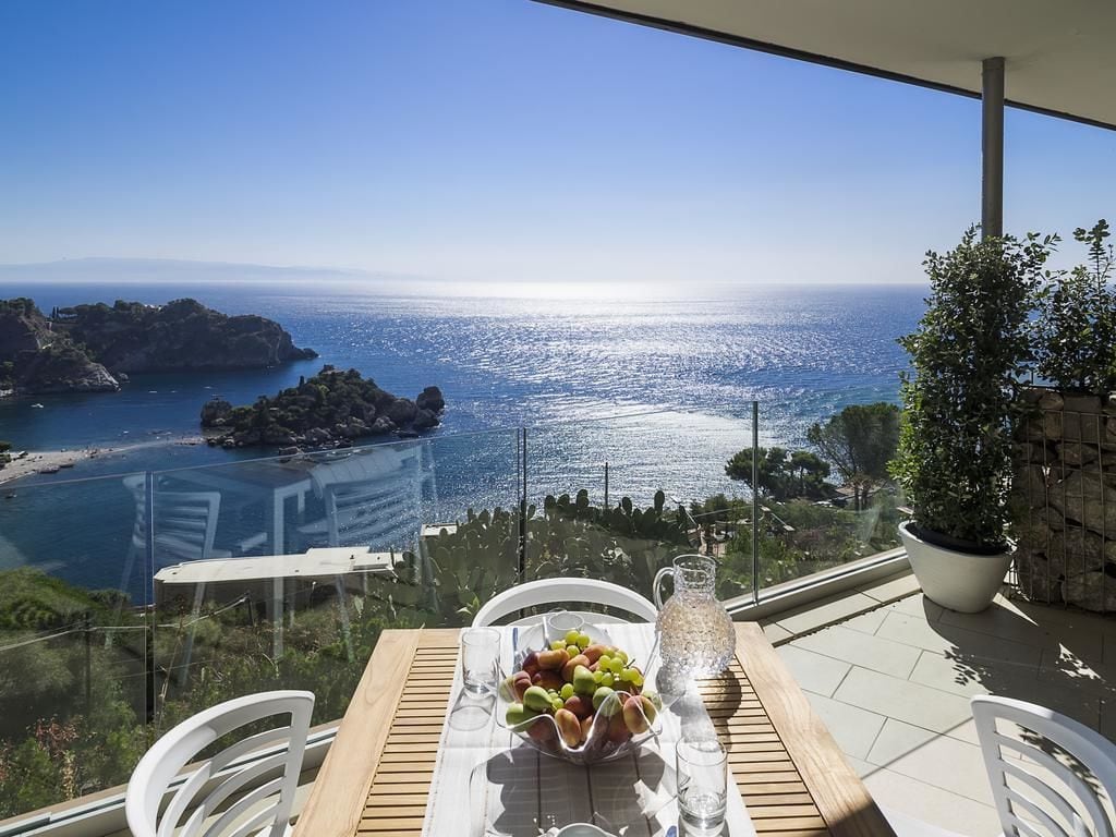 A table with a fruit bowl and pitcher, overlooking a coastal view with a clear sky.