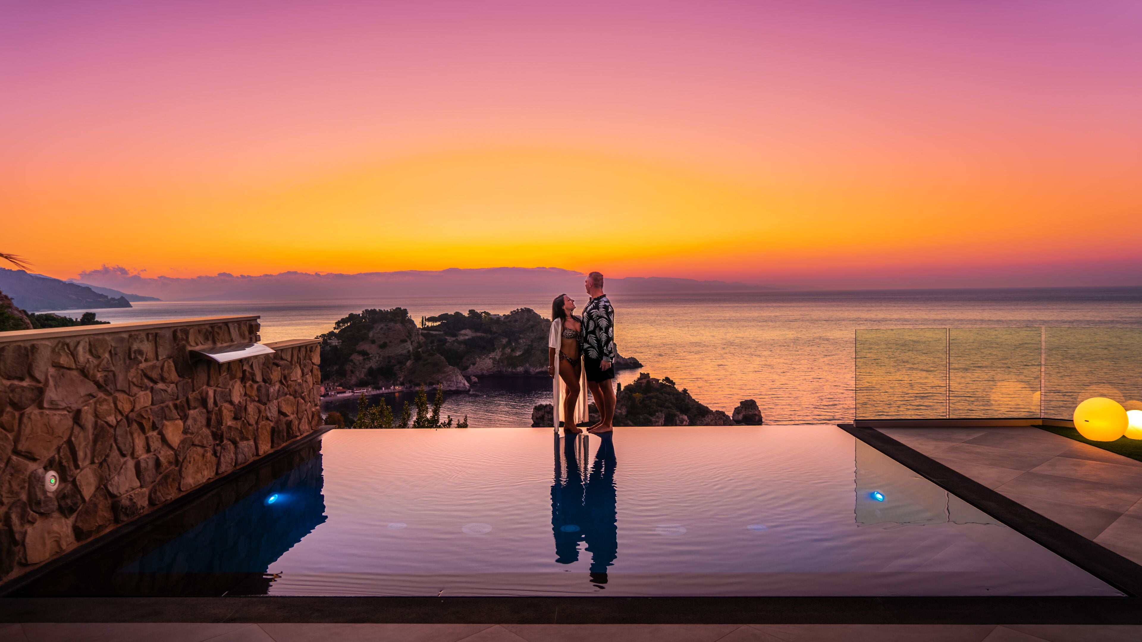 A couple standing on a infinity pool with a sunset view.