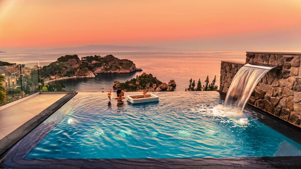 A infinity pool with a waterfall feature, a boat, and a view of a coastline at sunset. - Isola Bella Infinity Suites (Taormina)