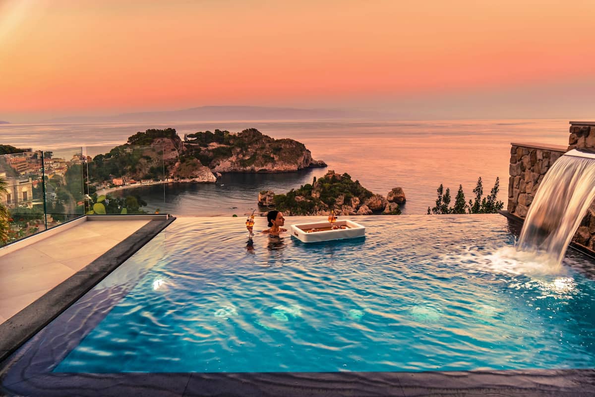 Una piscina infinita con cascada, un bote y vistas a la costa al atardecer.