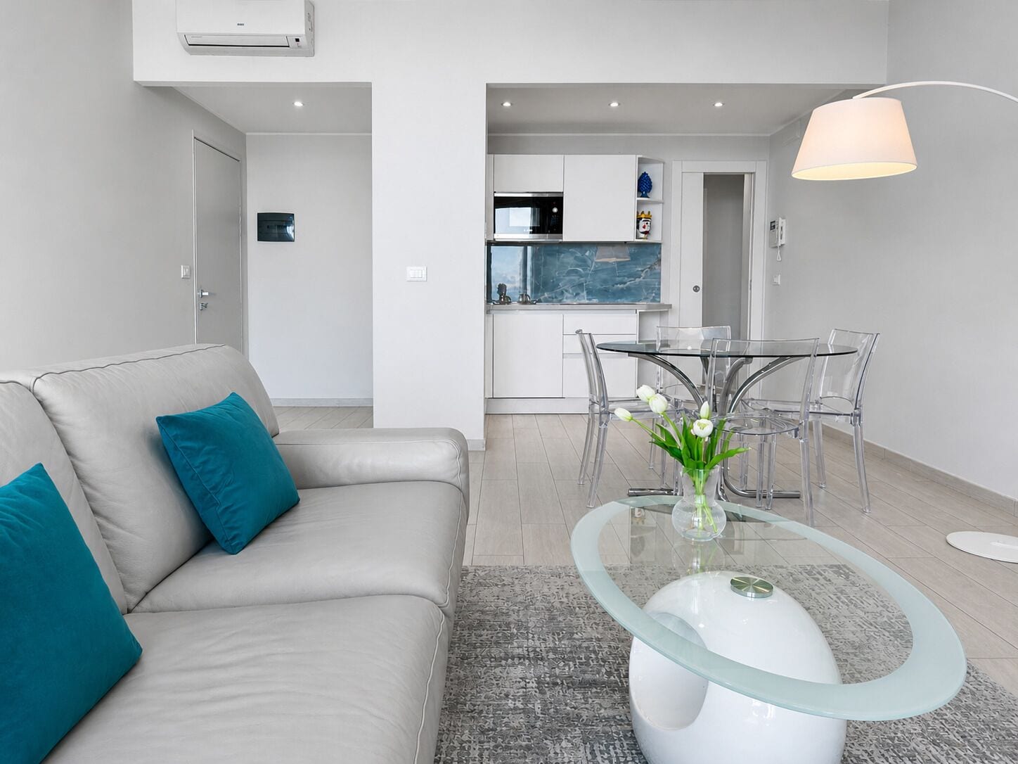A modern living room with a glass coffee table, a beige sofa with teal cushions, and a dining area with a glass table and chairs.