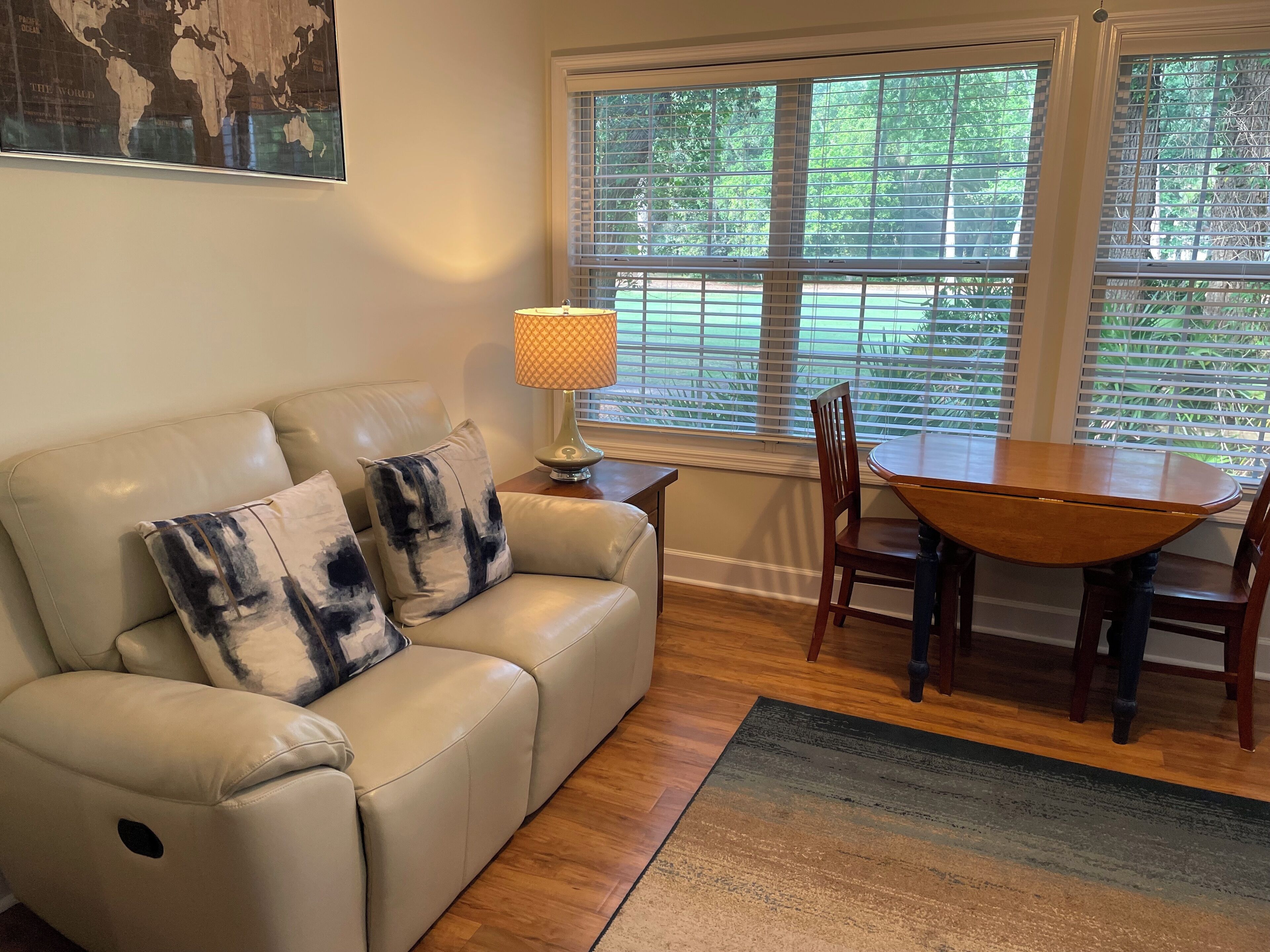 Un salon avec un canapé en cuir beige, deux coussins décoratifs à motifs, une table à manger en bois avec quatre chaises, un lampadaire et une grande fenêtre avec des stores.
