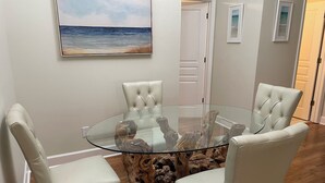 A dining area with a glass table surrounded by white upholstered chairs, a wooden trunk centerpiece, and a painting of a beach scene on the wall. - Grace Meadows: 3BR/3BA, close To Westin, Beach, Golf & Tennis (Hilton Head Island)