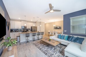 Une cuisine moderne avec des meubles blancs, un îlot central doté de tabourets de bar, un coin repas avec un canapé blanc et des coussins bleus, et un ventilateur de plafond.