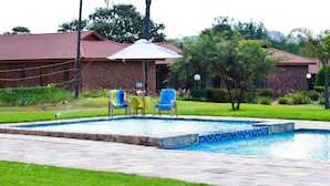 A swimming pool with a tiled edge, surrounded by a grassy area and a brick building with a red roof. - Zororo lodge (Polokwane)
