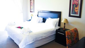 A neatly made bed with white linens, a nightstand with a lamp, and a chair with a floral pattern. - Zororo lodge (Polokwane)