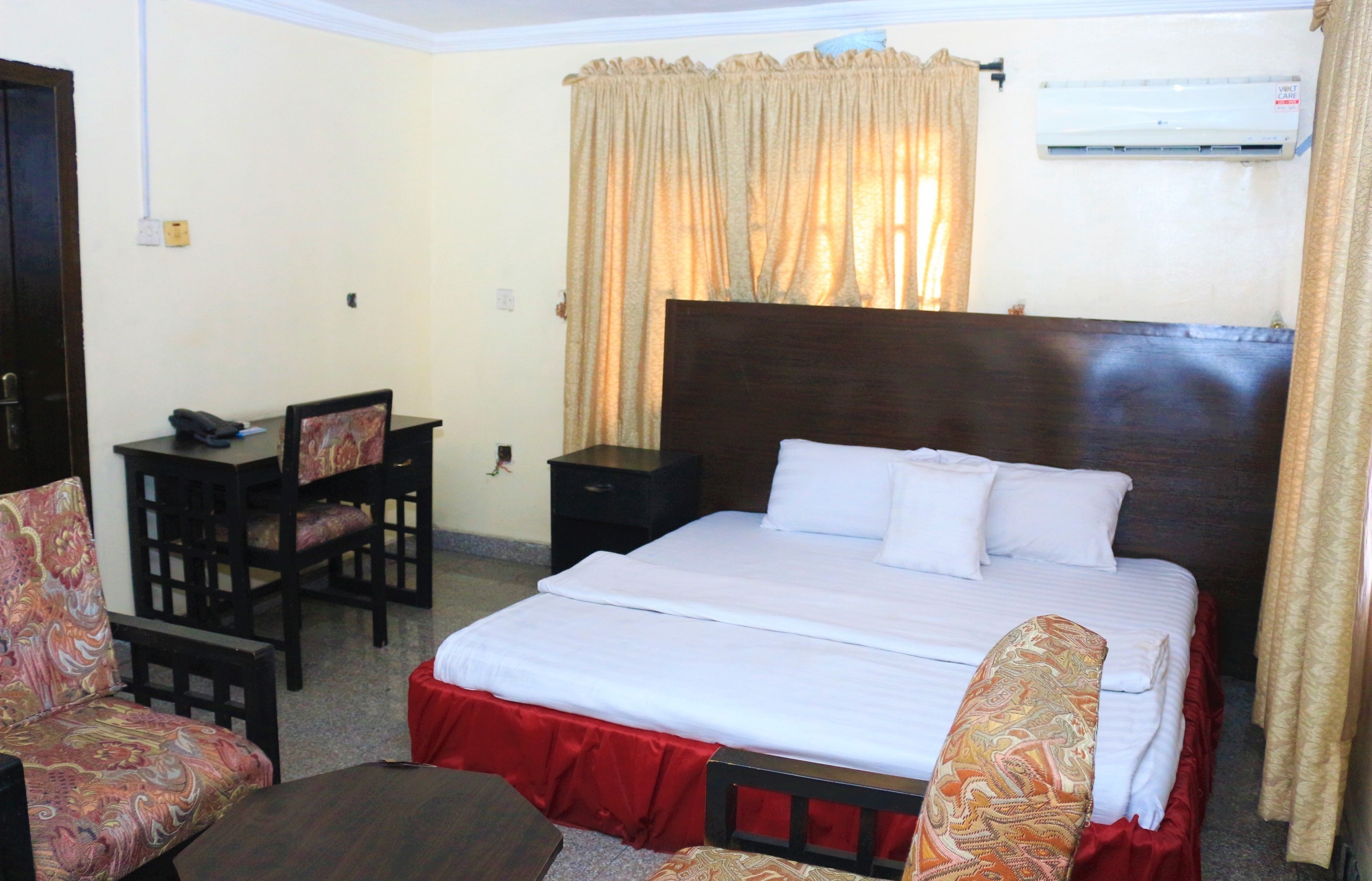 A hotel room with a bed, a desk, a chair, and an air conditioning unit.