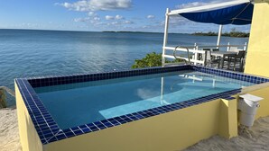 Una piscina amb rajoles blaves, envoltada de sorra, amb vista al mar i una tanca blanca.