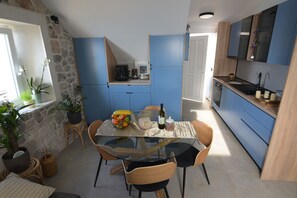 A compact kitchen with blue cabinets, a glass dining table, and a stone accent wall. - Perfect Beachfront Holiday In a Secluded Traditional Stone House With Pool (Mimice)