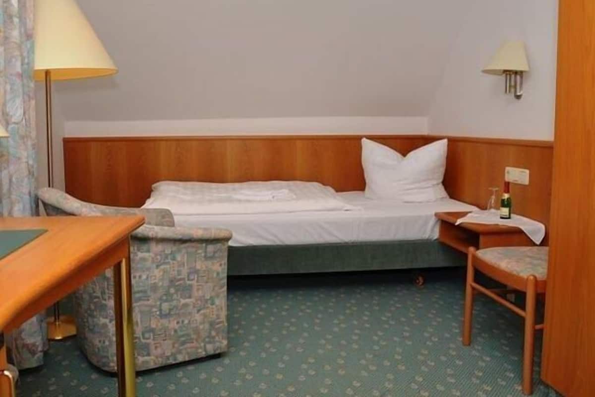 Una habitación de hotel pequeña y sencilla, con una cama individual, una silla, una mesita y una lámpara.