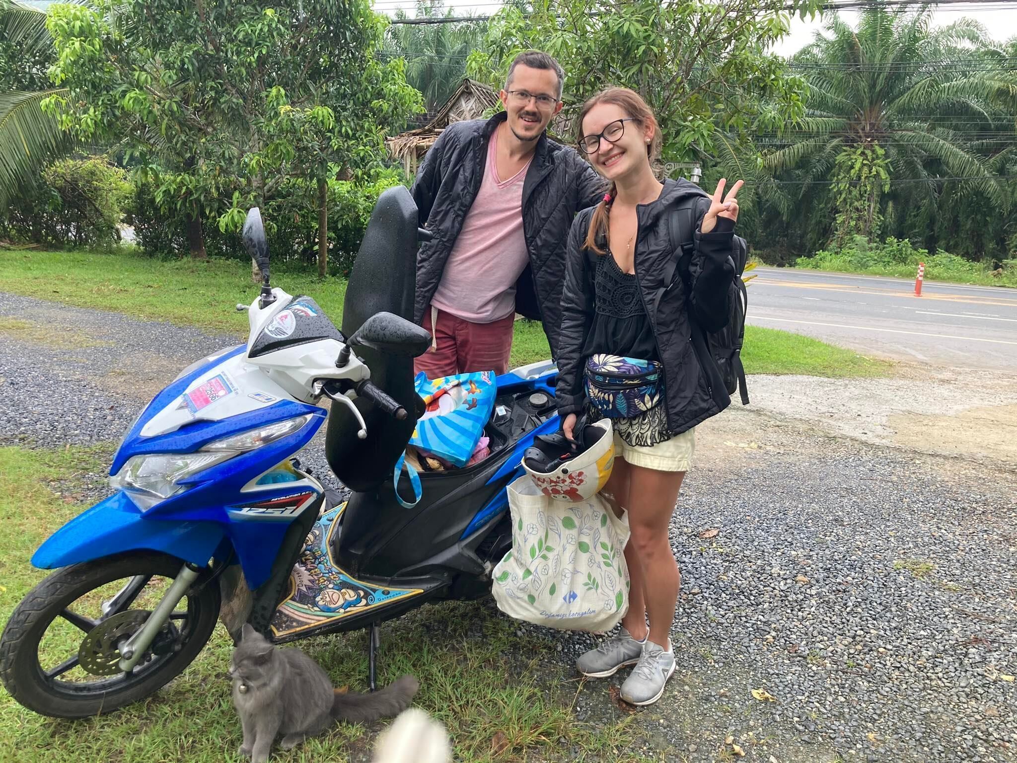 A blue scooter with a decorative seat and a bag on it, parked on a gravel path with greenery in the background.