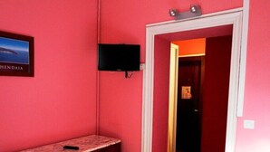 A room with pink walls, a wooden dresser with drawers, a flat-screen TV mounted on the wall, and a door leading to another room. - Hôtel Bellevue (Hendaye)