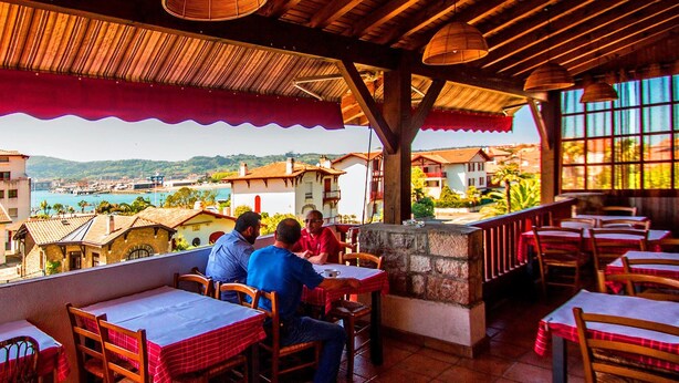An outdoor restaurant with wooden tables and chairs, a stone fireplace, and a view of the town and hills in the background. - Hôtel Bellevue (Hendaye)