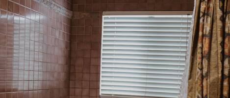 A bathroom with a bathtub, a showerhead, and a window with blinds.