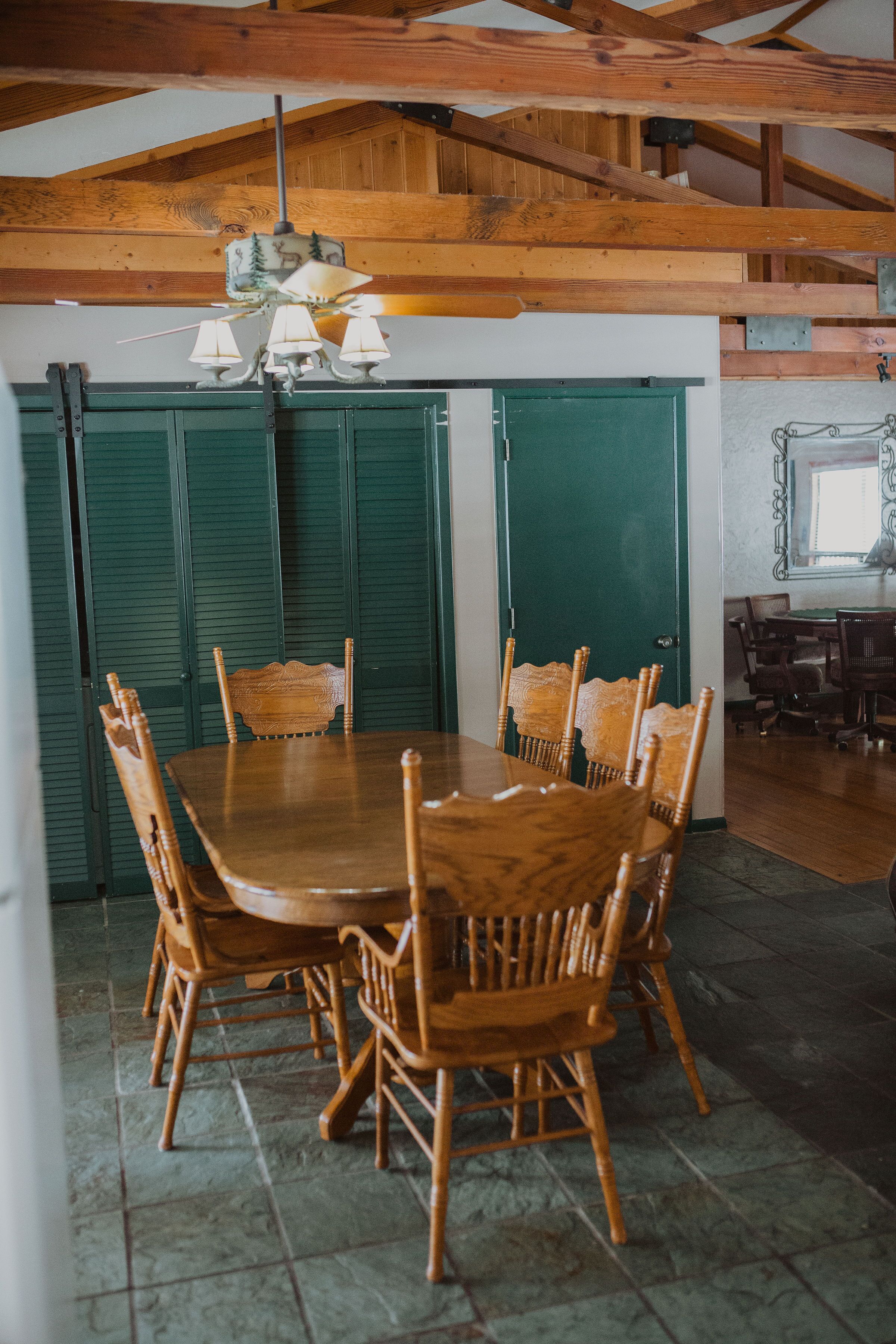 A wooden dining table with six chairs, a chandelier, and green shutters in a room with a high ceiling and exposed beams.