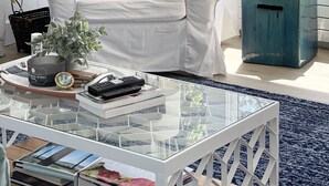 Una poltrona bianca con cuscini decorati, un tavolino da caffè in vetro con libri e una pianta, e vista mare attraverso un'ampia finestra.