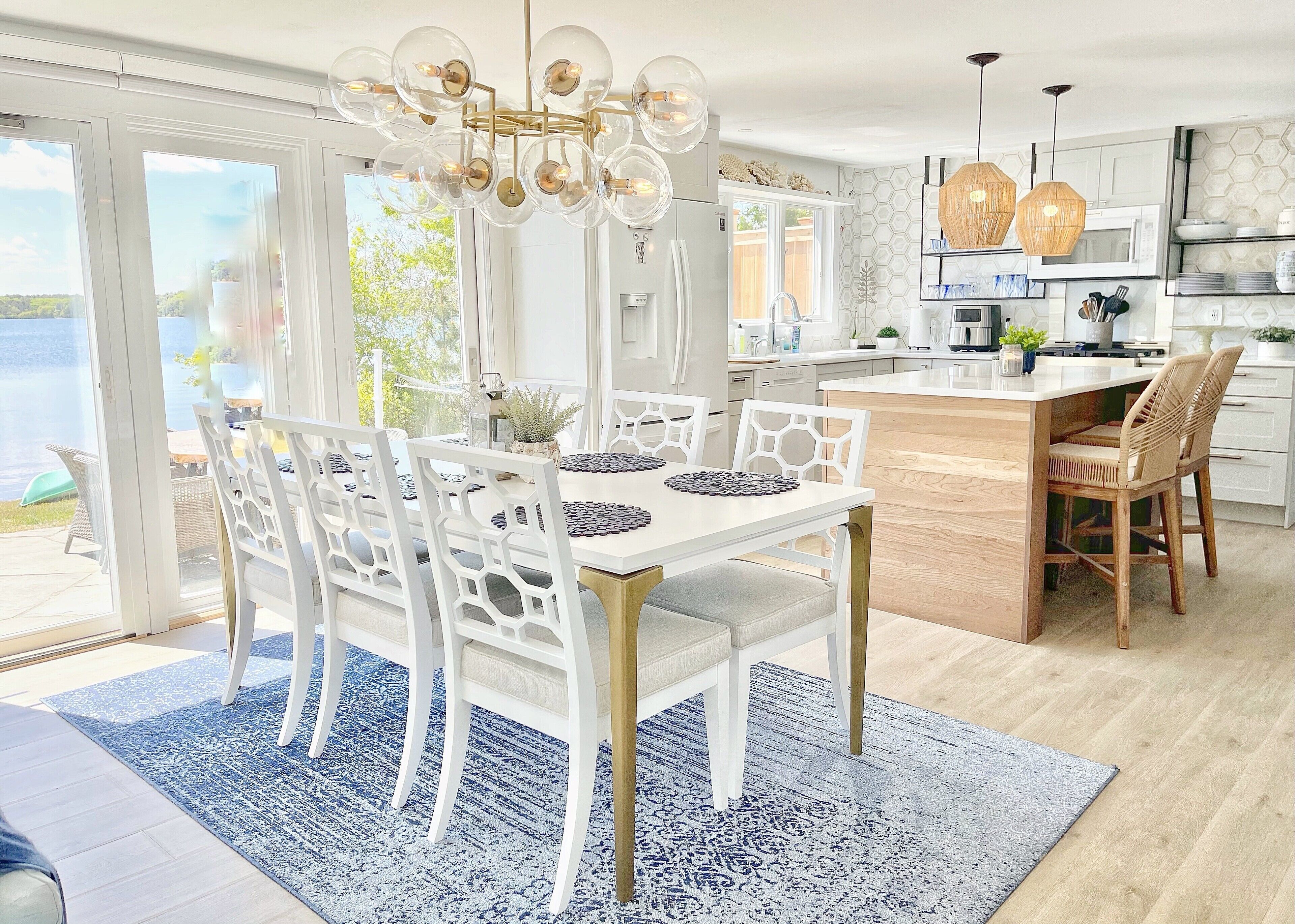 A modern kitchen with white cabinetry, a central island, and a dining area with white chairs and a blue patterned rug.