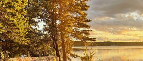 A wicker sofa with striped cushions and a decorative pillow, positioned on a stone patio overlooking a calm lake, with trees and a cloudy sky in the background.