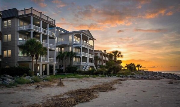Een hotel aan het strand met meerdere balkons, een zandstrand ervoor en een ondergaande zon op de achtergrond.