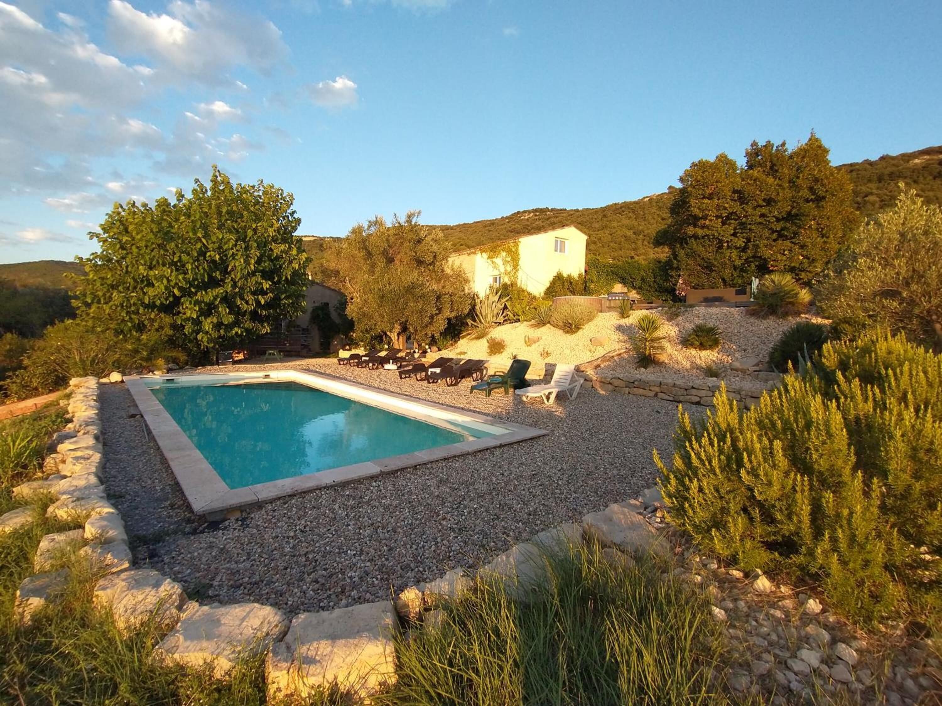 A pool surrounded by rocks and plants, with lounge chairs and a building in the background.