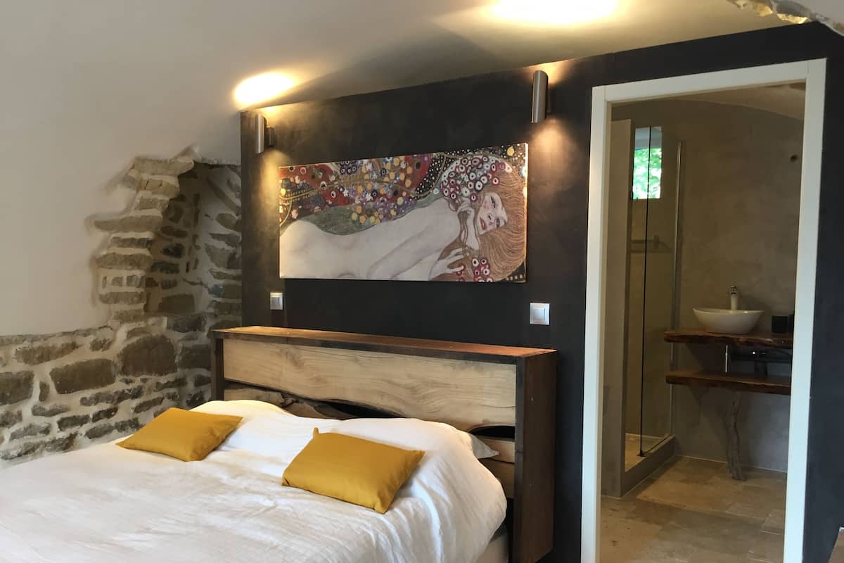 Une chambre à coucher avec un lit, une tête de lit en bois, un mur en pierre, un tableau, et une salle de bain avec un lavabo.