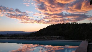 A swimming pool reflecting a sunset sky with clouds, surrounded by a stone wall and a forested hillside. - Les Agaves (Grospierres)