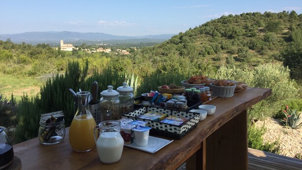 A breakfast table with various food items and drinks, overlooking a scenic landscape with hills and a distant building. - Les Agaves (Grospierres)