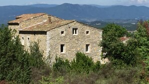 A stone building with a sloped roof, surrounded by greenery and mountains in the background. - Les Agaves (Grospierres)