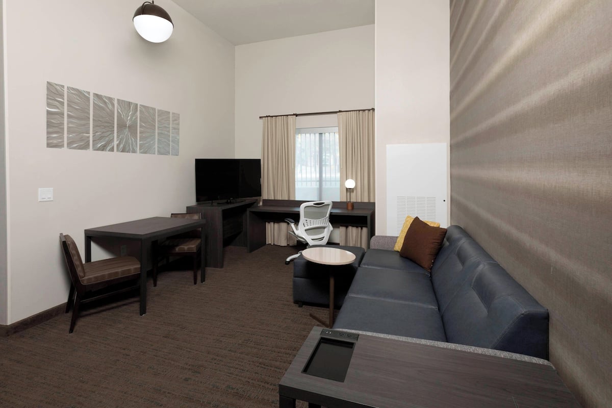 A hotel room with a dark brown carpet, a blue sofa, a wooden desk with a chair, a flat-screen TV, and a window with curtains.