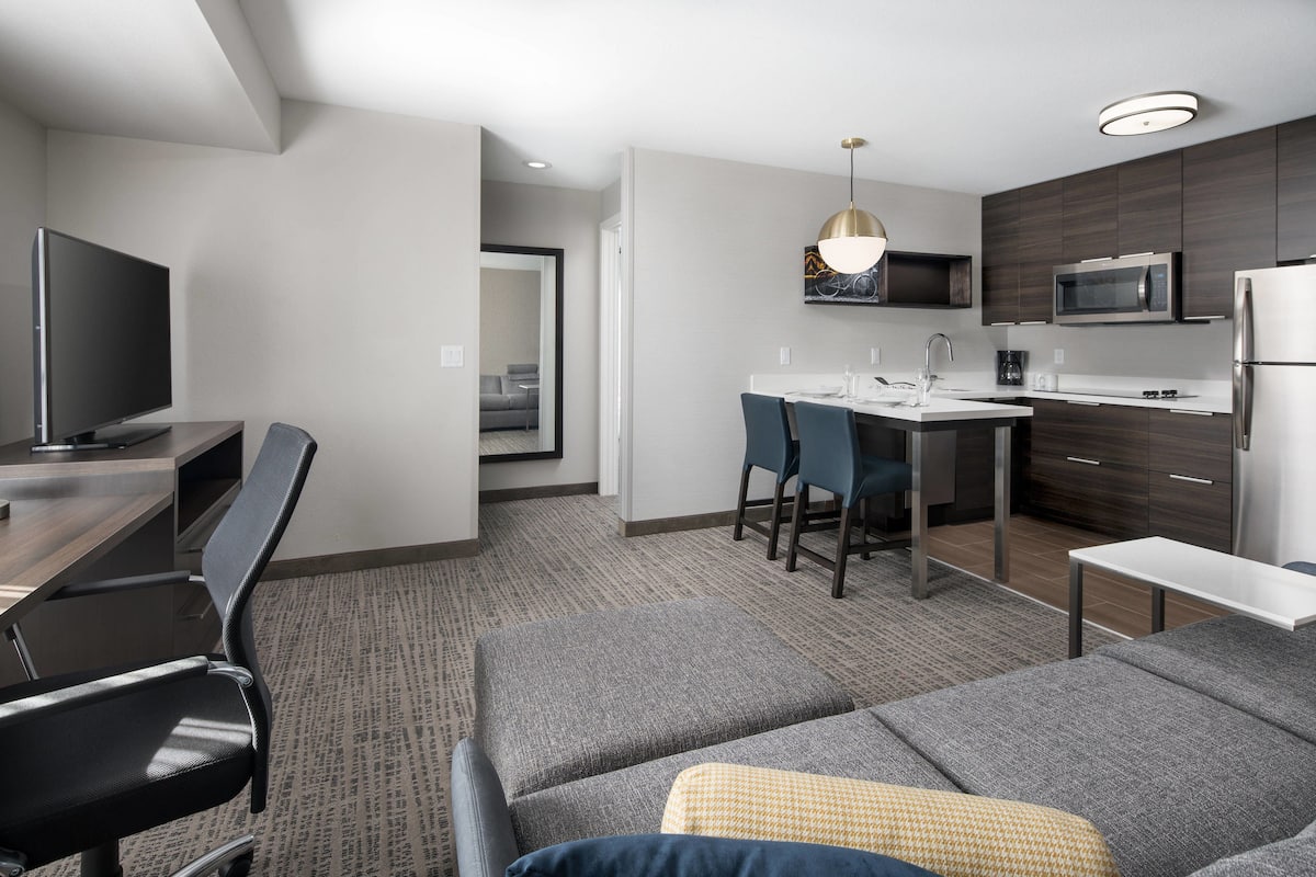 A modern hotel room with a sofa, a dining area with chairs, a TV, and a kitchen with a microwave and refrigerator.