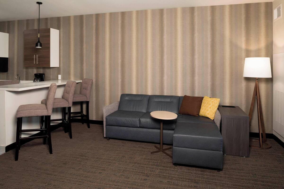 A modern hotel room with a grey sectional sofa, a small round table, a desk with a chair, and a lamp.