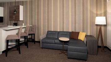 A modern hotel room with a grey sectional sofa, a small round table, a desk with a chair, and a lamp.