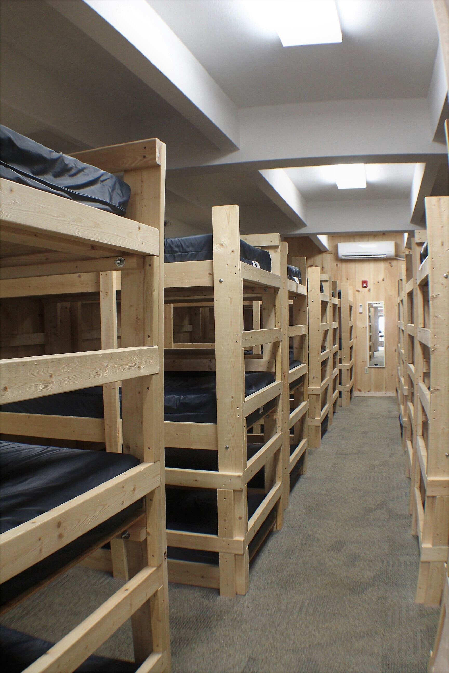 A corridor with bunk beds arranged on either side, a carpeted floor, and an air conditioning unit on the wall.