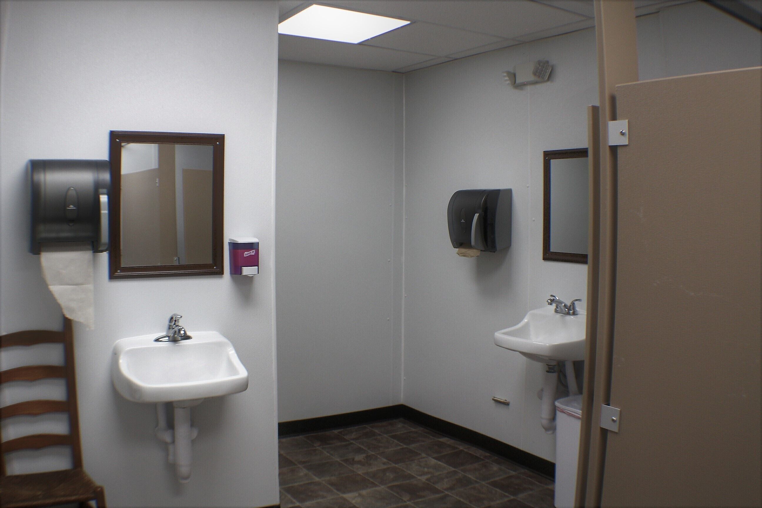 Une petite salle de bain avec deux lavabos, un miroir, un distributeur de serviettes et une chaise.