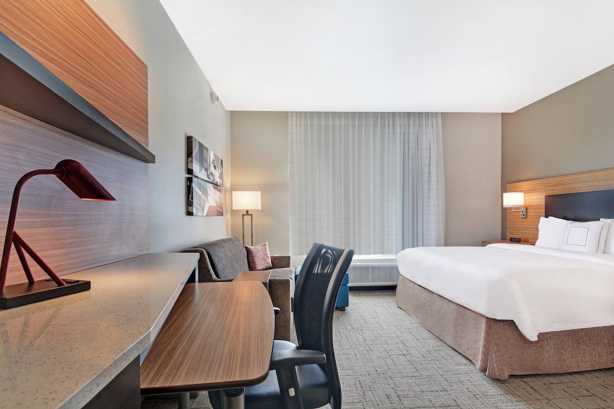 Habitación de hotel con cama, escritorio, silla y un gran ventanal con cortinas.