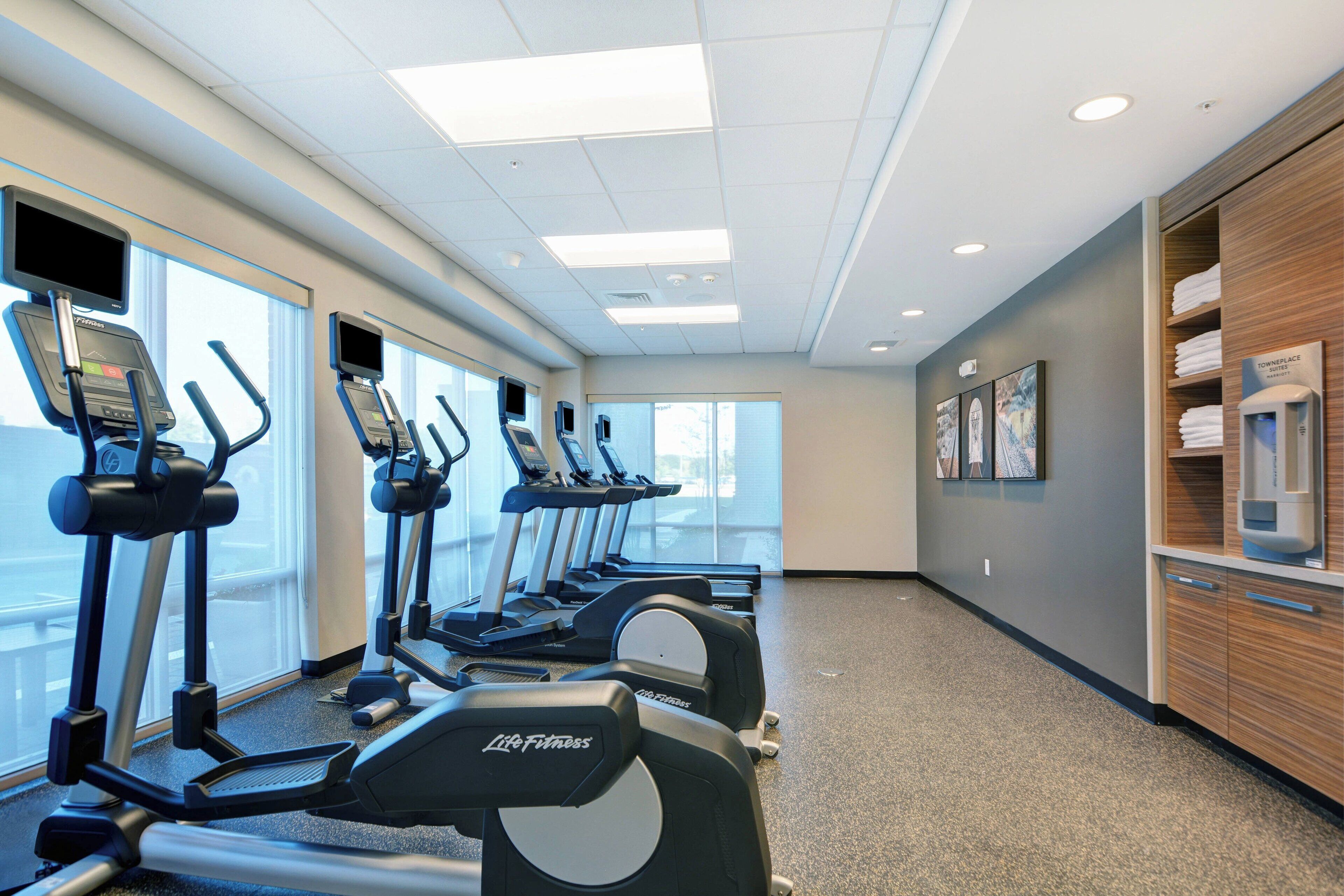 Un gimnasio con cintas de correr y máquinas elípticas, amplias ventanas y una zona de recepción con mostrador y dispensador de agua.